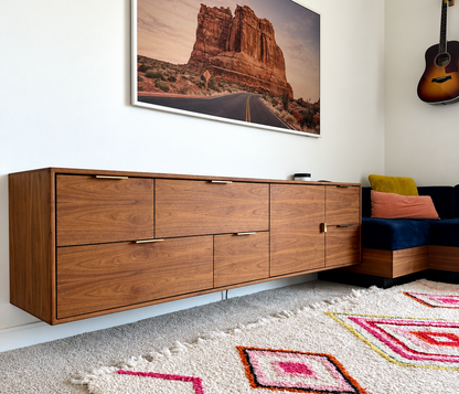 A mid-century modern wooden credenza or sideboard with six drawers, shown in a styled living room setting with a large framed landscape print on the wall, a patterned rug, and a sectional sofa.