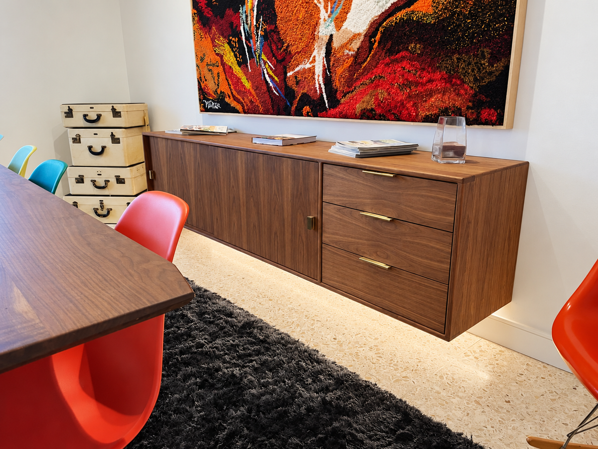 A mid-century modern wooden credenza or sideboard with a wood-grain finish, featuring a cabinet door on the left and two drawers on the right with gold handles. It is styled in a room with a colorful abstract painting on the wall, vintage suitcase storage, and orange chairs.